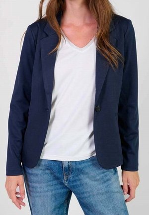 Frau trägt einen marineblauen Blazer, weißes V-Ausschnitt-Shirt und blaue Jeans, die Hände entspannt an den Seiten.