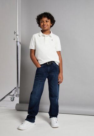 Polo bianco, jeans blu scuro e sneakers bianche. Giovane con capelli ricci si erge con sicurezza, con le mani in tasca, su uno sfondo semplice.