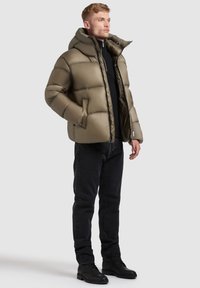 Olive-green puffer jacket with quilted design, high collar, and zip closure. Worn over a black sweater and paired with black jeans and boots.