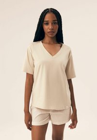 Beżowa koszulka polo z dekoltem w serek i krótkimi rękawami, zestawiona z jasnymi szortami w paski, charakteryzującymi się luźnym krojem i gładką fakturą. Metka widoczna na koszulce.