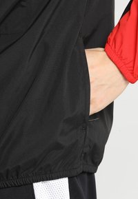 Zwarte windbreaker jas met rode accenten, met een gestructureerd oppervlak, elastische manchetten en een zak op de taille. De zoom is afgerimpeld.