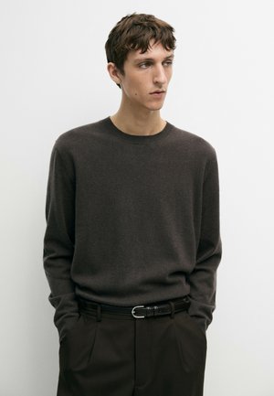 Jeune homme aux cheveux courts et bruns portant un pull marron foncé et une ceinture noire, debout les mains dans les poches, regardant vers la droite sur un fond uni.