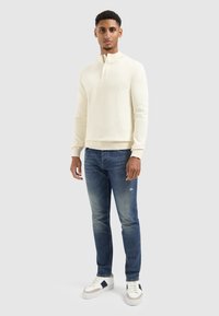 Crème gebreide sweater met hoge kraag, gecombineerd met donkerblauwe slim-fit jeans en witte sneakers met marineblauwe accenten.