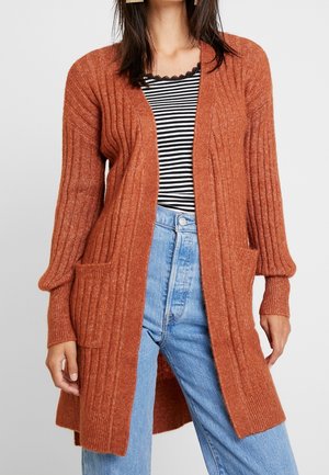 Femme portant un cardigan côtelé couleur rouille sur un haut rayé noir et blanc et un jean taille haute bleu clair.