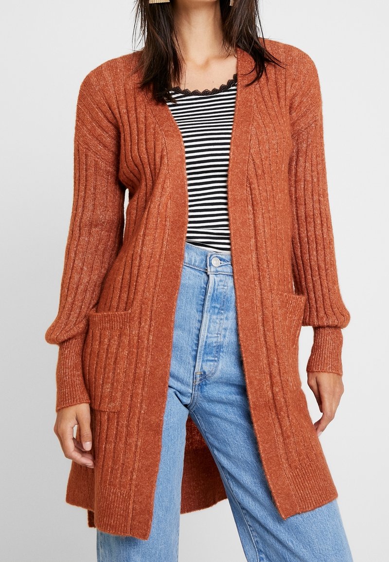 Femme portant un cardigan côtelé couleur rouille sur un haut rayé noir et blanc et un jean taille haute bleu clair.