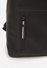 Zaino nero con superficie texturizzata, tasca laterale con zip nera e un tirazip decorativo con righe rosse, bianche e blu.