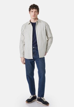 Młody mężczyzna stoi, ubrany w beżową koszulę w paski na guziki, pod którą ma granatowy t-shirt, niebieskie jeansy i czarne trampki z białymi sznurowadłami.