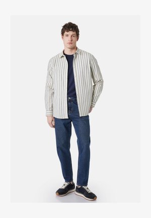 Giovane uomo in piedi che indossa una camicia beige a righe con bottoni sopra una t-shirt blu navy, jeans blu e sneakers nere con lacci bianchi.