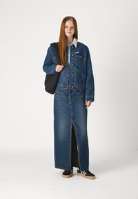 Τζάκετ από τζιν με υφασμάτινο γιακά, συνδυασμένο με μια μακριά τζιν φούστα με σχισμή μπροστά. Μαύρη τσάντα και sneakers με λευκές και μωβ ρίγες.