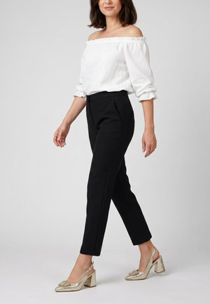 Žena kráčí v bílé blůze s odhalenými rameny, černých kalhotách na míru a zlatých botách na blokovém podpatku se zapínáním.