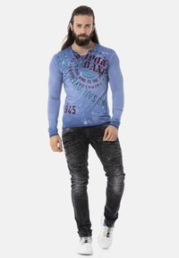 Blaues Langarmhemd mit grafischen Drucken, kombiniert mit schwarzen, strukturierten Cargo-Jeans. Das Model trägt weiße Sneakers mit bunten Akzenten.