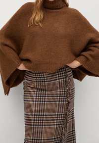 Maglione a collo alto lavorato a maglia marrone con maniche larghe, abbinato a una gonna a quadri al ginocchio con toni marroni, blu e rossi.