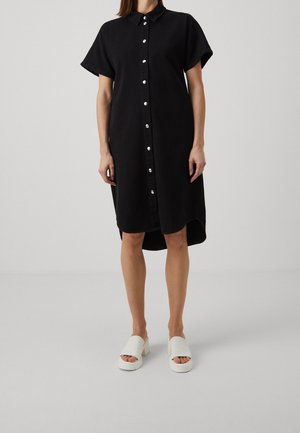 Femme portant une robe-chemise noire à manches courtes avec boutons-pression sur le devant et des sandales blanches à talons blocs ouverts sur un fond uni.