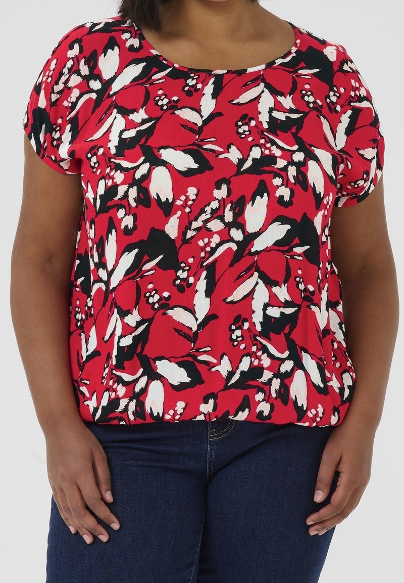 Femme portant un haut rouge à manches courtes avec un motif floral blanc et noir et un jean bleu foncé, debout les mains détendues le long du corps.