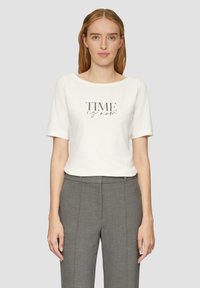 T-shirt blanc à manches courtes avec le texte "TIME is now" en gris. Assorti à un pantalon tailleur gris en tissu lisse.