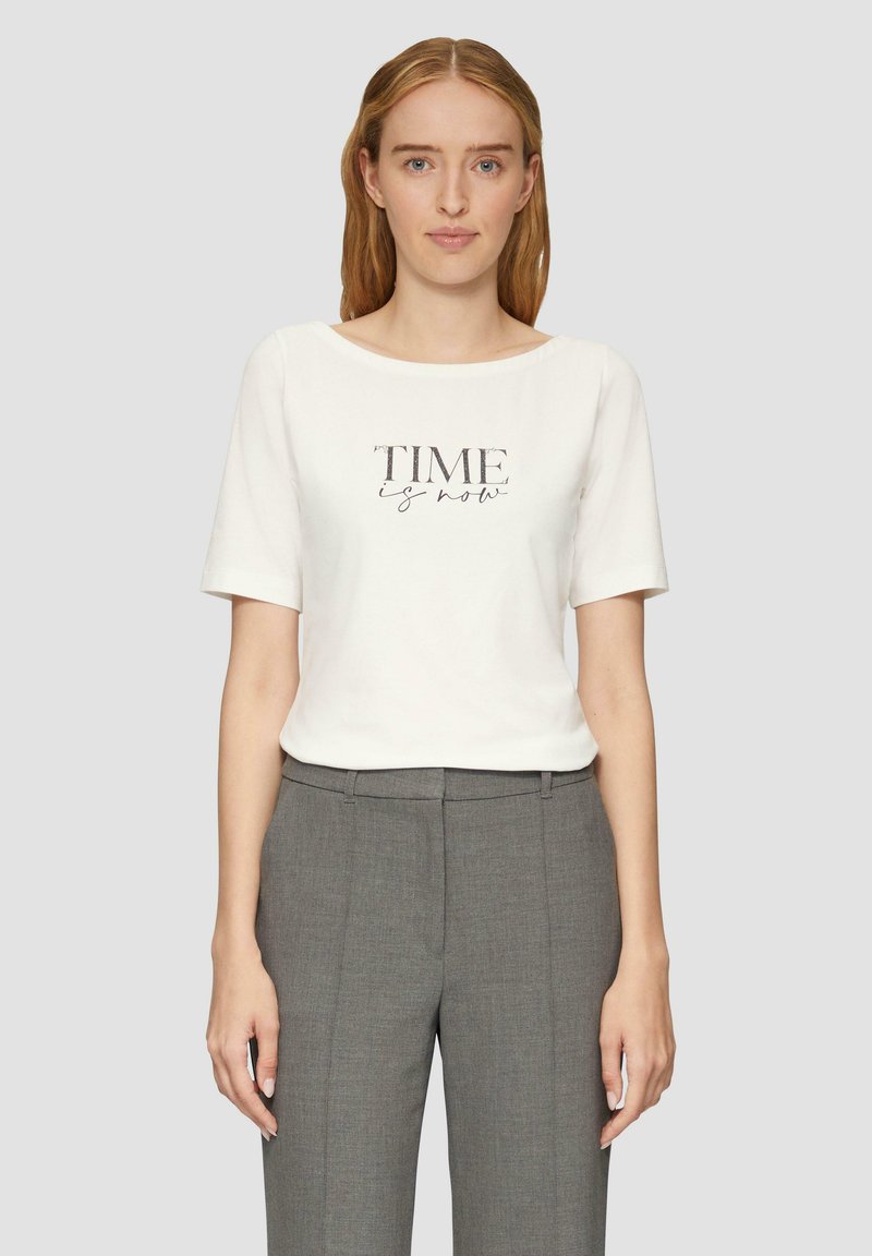 T-shirt blanc à manches courtes avec le texte "TIME is now" en gris. Assorti à un pantalon tailleur gris en tissu lisse.