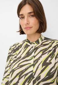 Colbert blouse met een lichtbeige basis en groene en donkerbruine zebrapatronen. Zachte stof met geplooide schouderdetails.