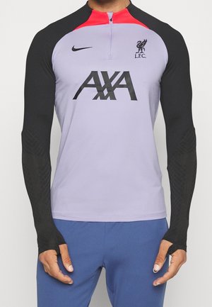 Homme portant un haut de sport à manches longues avec des manches noires, un torse violet, un col rouge, des logos Nike et Liverpool FC, ainsi qu'un logo de sponsor AXA, associé à un pantalon bleu.