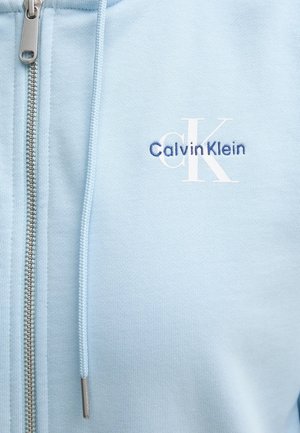 Světle modrá mikina na zip vyrobená z jemné tkaniny. Obsahuje stříbrný zip a vyšívané logo "Calvin Klein" v námořnické modré a bílé barvě.