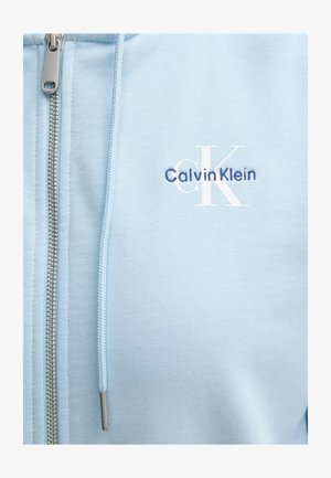 Světle modrá mikina na zip vyrobená z jemné tkaniny. Obsahuje stříbrný zip a vyšívané logo "Calvin Klein" v námořnické modré a bílé barvě.