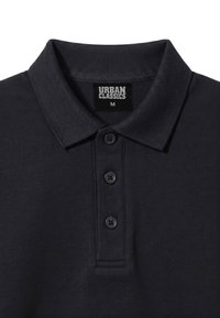 Polo noir avec col côtelé et trois boutons, avec une étiquette indiquant "Urban Classics" et la taille medium à l'intérieur de l'encolure.