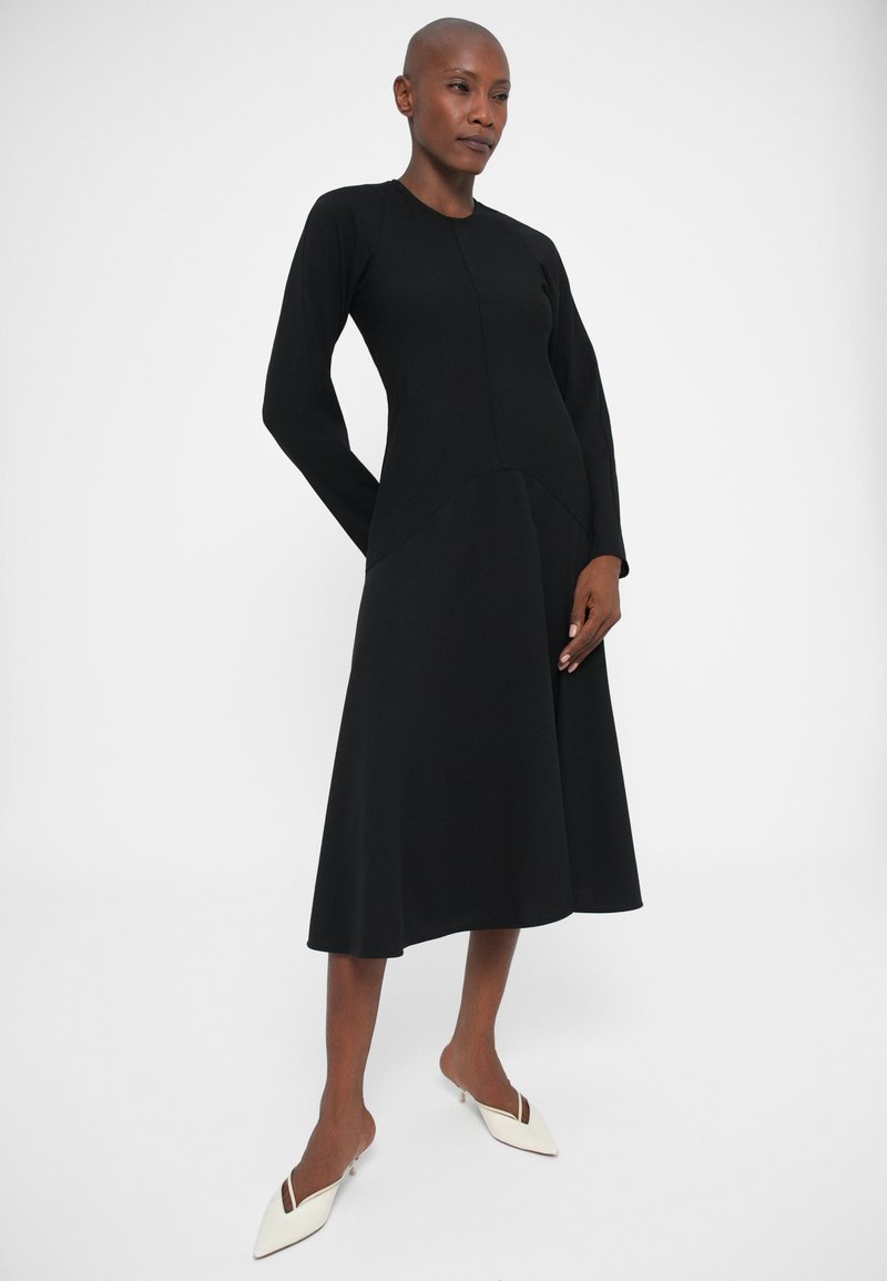 Femme portant une longue robe noire à manches longues et des mules à talons kitten blancs à bout pointu, se tenant devant un fond blanc uni.