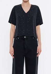 Černý krátký rukáv top s V-výstřihem, zdobený malými barevnými perlami po celém povrchu. Spárováno s černými džínovými kalhotami s vysokým pasem.