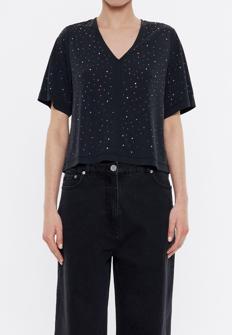 Černý krátký rukáv top s V-výstřihem, zdobený malými barevnými perlami po celém povrchu. Spárováno s černými džínovými kalhotami s vysokým pasem.