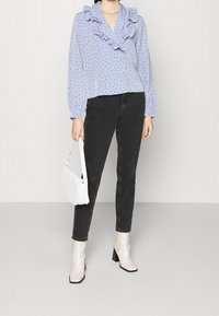 Blusa azzurra con pois neri e scollatura arricciata, abbinata a jeans neri e borsa a tracolla bianca trapuntata. Stivaletti bianchi alla caviglia completano il look.