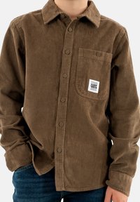 Camisa de pana marrón con cuello abotonado, bolsillo frontal y textura acanalada visible. Incluye una etiqueta en el bolsillo.