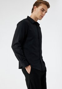 Czarna, długą rękawą koszula z gładkiego materiału, z kołnierzykiem, guzikami i mankietami. W zestawie z dopasowanymi czarnymi spodniami, widoczna boczna kieszeń.