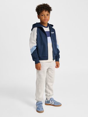 Dreng stående, iført navy og grå lynlåshættetrøje, stribet skjorte, lysegrå joggingbukser og blå sneakers mod ensfarvet baggrund.