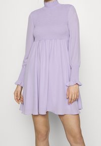 Levandulové šaty s dlouhým rukávem, s nabíraným živůtkem, vysokým výstřihem a rozšířenou sukní. Měkká látka s nařasenými manžetami rukávů a splývavým lemem.