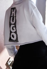 Bílá zkrácená mikina s černým grafickým nápisem "HUGO" na zádech. Hladká textura, volný střih. Kombinována s černými kalhotami s vysokým pasem.