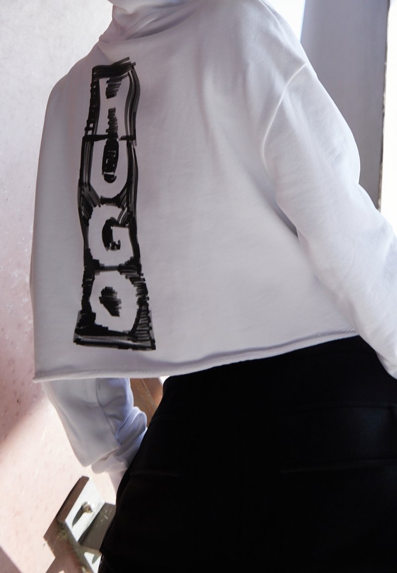 Bílá zkrácená mikina s černým grafickým nápisem "HUGO" na zádech. Hladká textura, volný střih. Kombinována s černými kalhotami s vysokým pasem.