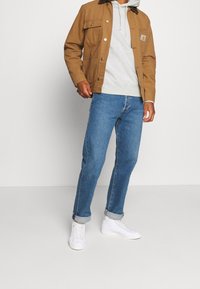 Brun canvasjacka med två bröstfickor över en ljusgrå hoodie. Blå denimjeans, uppvikta vid anklarna, kombinerade med vita sneakers.