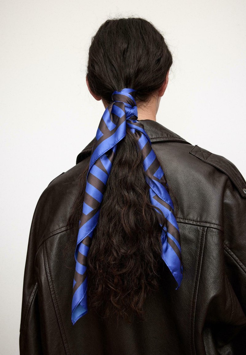 Femme aux longs cheveux bouclés foncés attachés avec un foulard rayé bleu et noir, portant une veste en cuir marron foncé, vue de dos.
