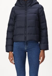 Persona che indossa una giacca imbottita nera con cappuccio e jeans di denim blu, in piedi con le braccia rilassate lungo i lati.