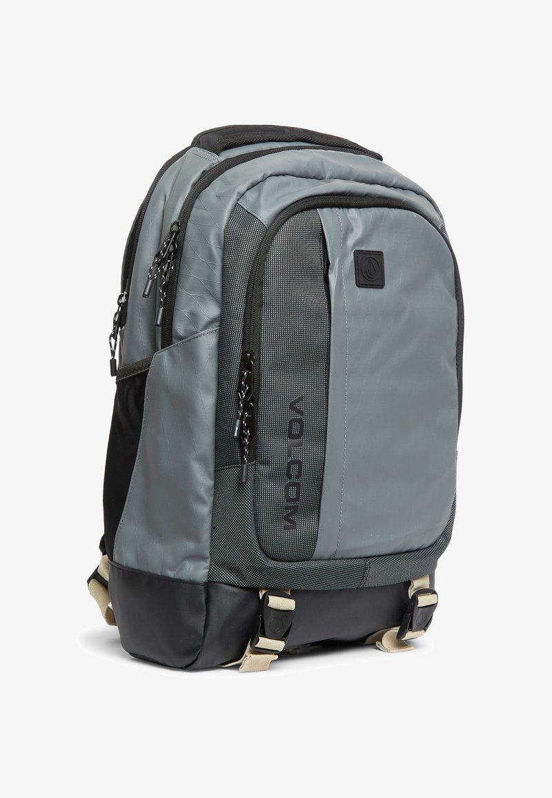 Sac à dos Volcom gris et noir avec plusieurs compartiments zippés et des bretelles beiges réglables en bas.