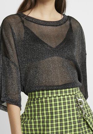 Haut noir transparent avec des taches argentées, manches larges, superposé sur un débardeur noir. Associé à une jupe à carreaux verts et noirs avec un détail en crochet métallique.