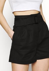 Shorts neri a vita alta con una larga cintura dotata di una fibbia rettangolare, fronte plissettato e tasche laterali, realizzati in un tessuto liscio.
