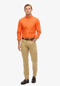 Pomarańczowa koszula zapinana na guziki, khaki spodnie, brązowy pleciony pasek i ciemne loafersy. Koszula ma kołnierzyk i jest włożona w spodnie.
