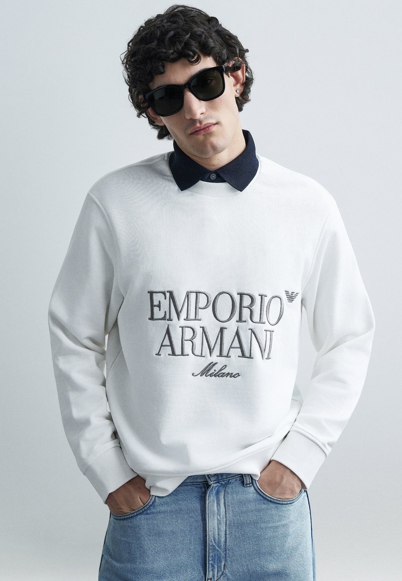 Sweatshirt branca com gola azul-marinho, com um logótipo bordado "Emporio Armani Milano". Usada com óculos de sol pretos. Corte standard.