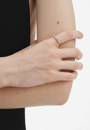 Mano con anillo incrustado de diamantes descansando sobre un brazo flexionado, lunares visibles en la piel, sobre un fondo blanco con ropa negra.