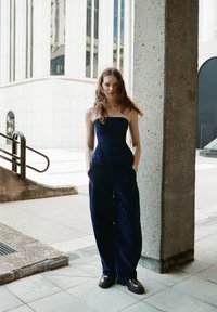 Temno modra brezramna peplum bluzo v kombinaciji z širokimi hlačami, ki imajo mehko teksturo. Model stoji ob betonski strukturi v urbanem okolju.