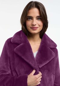 Abrigo de piel sintética púrpura con un gran collar, superficie texturizada y cierre frontal, llevado sobre una blusa gris claro.
