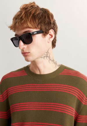 Gafas de sol rectangulares negras con lentes oscuras. Llevadas con un suéter verde texturizado que presenta franjas horizontales rojas. Tatuaje notable en el cuello.