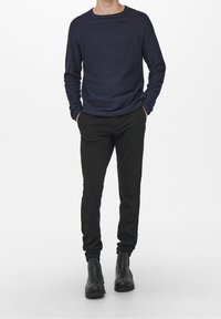 Jeune personne portant une chemise à manches longues bleu marine, pantalon noir ajusté, bottes noires, les mains dans les poches, debout devant un fond blanc uni.