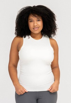Woman with curly hair wearing a white sleeveless top and gray leggings, standing with hands near hips against a plain background.