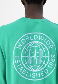 Green cotton sweatshirt featuring a large white graphic with the text "WORLDWIDE GAP ESTABLISHED 1969" and a globe design on the back.